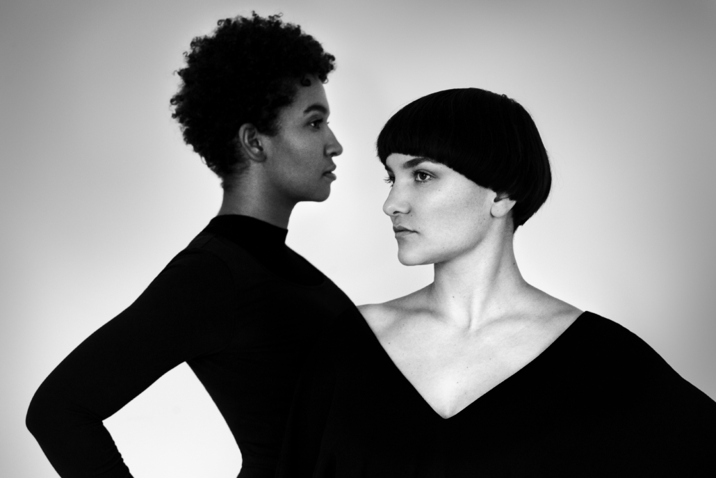 Schwarz-weiß-Bild von zwei Frauen mit dunklen Haaren, eine mit kürzeren Haaren, eine mit kurzen lockigen Haaren in schwarzen Oberteilen, die ineinander übergehen. Zugewandt, Blick aneinander vorbei