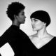 Schwarz-weiß-Bild von zwei Frauen mit dunklen Haaren, eine mit kürzeren Haaren, eine mit kurzen lockigen Haaren in schwarzen Oberteilen, die ineinander übergehen. Zugewandt, Blick aneinander vorbei
