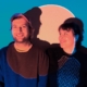 Junger Mann mit Käppi und junge Frau mit kürzeren Haaren vor blauer Wand, werden von einem warmen runden Scheinwerferlicht beschienen.