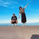 Ein Mann und eine Frau befinden sich draußen an einem Gewässer und stehen auf einer betonierten Fläche. Der Mann hockt in sportlicher Kleidung mit T-Shirt, Sneakers und einer Flasche auf dem Boden. Die Frau balanciert mit ausgebreitetem Arm, trägt eine gemusterte Bluse, einen dunklen Rock und helle Turnschuhe. Im Hintergrund ist das ruhige, blaue Meer und ein weiter, fast wolkenloser Himmel zu sehen.