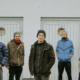 Fünf junge Männer stehen in Winterbekleidung vor einer hellen Wand mit hellen Türen und kleinen runden Fenstern