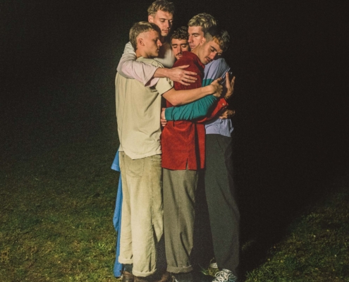 Das Bild zeigt fünf junge Menschen, die sich auf einer Wiese oder einer Freifläche im Dunkeln eng umarmen. Sie stehen dicht beieinander und legen ihre Arme umeinander, sodass ein Gefühl von Nähe und Gemeinschaft entsteht. Die Gruppe ist von vorne beleuchtet, während der Hintergrund fast vollständig schwarz ist und nur der Boden leicht sichtbar bleibt. Die Personen tragen unterschiedliche, eher legere Kleidung: Hemden, Jacken und Hosen sowie Sneakers.