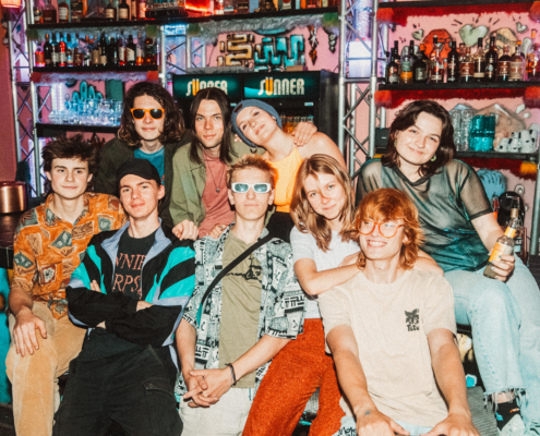 Bild im Blitzlicht-Stil: Das Bild zeigt eine Gruppe junger Menschen, die in einer bunten Bar oder Lounge zusammen sitzen. Im Vordergrund sitzen vier Personen auf einer Bank, während fünf weitere Personen hinter ihnen stehen oder auf der Bar sitzen. Die Personen wirken entspannt, einige unterhalten sich und eine Person hält eine Flasche Getränk in der Hand. Im Hintergrund sind Regale mit vielen Flaschen und Gläsern zu sehen, bunt beleuchtet und von Graffiti-Dekorationen umgeben. Die Stimmung wirkt ausgelassen und freundlich, als ob es sich um ein gemeinsames Treffen oder eine Feier handelt.